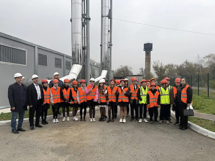 Группа людей в оранжевых жилетах и касках стоит на улице перед промышленным объектом с трубами и зданием. Погода пасмурная..